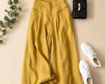 Puede incluir: Pantalones anchos de color amarillo mostaza con cintura elástica, pliegues delanteros y un detalle de canesú estructurado. Los pantalones se muestran junto a un bolso rectangular negro y un par de zapatillas blancas con cordones.