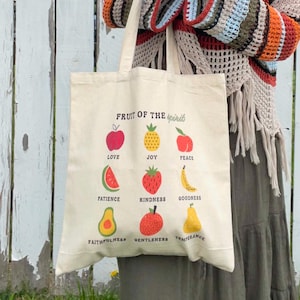 May include: Cream-colored tote bag featuring a colorful graphic of fruits with the words "Fruit of the Spirit" and related virtues. The fruits include an apple, pineapple, and pear. The bag has a simple design and is held in front of a white fence.