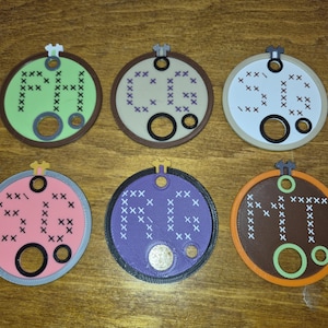 May include: Six circular, flat, 3D-printed embroidery hoop-shaped ornaments. Each features a unique color combination, a small hanger at the top, and two circular cutouts at the bottom. The center of each displays letters formed by cross-stitch patterns.
