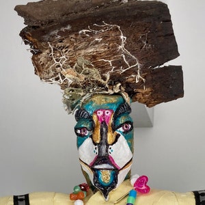 May include: A handcrafted sculpture featuring a painted face with teal, gold, and white accents, topped with a piece of weathered wood and natural lichen. The face has pink and black details, with a beaded necklace and a decorative pin.