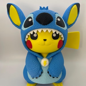 Puede incluir: Una figura impresa en 3D del personaje amarillo Pikachu vistiendo un disfraz azul de Stitch. La figura presenta grandes orejas azules con secciones internas amarillas, una capucha blanca con dientes, mejillas rojas, ojos negros y un emblema circular blanco en el pecho con un rayo amarillo.