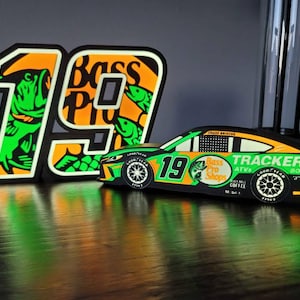 May include: A decorative set featuring a race car and the number 19, with the "Bass Pro Shops" logo. The car is black, green, and orange, with the number 19 on the side. The number 19 is also a separate piece, with a green fish design.