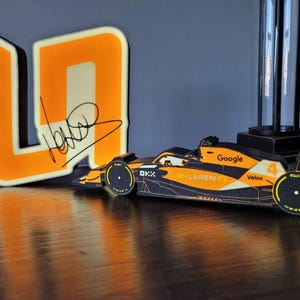 May include: An orange and black McLaren F1 race car model with the Google logo and the number 4. Beside it is a large orange and white number 4 with a signature. The items are on a dark wooden surface.