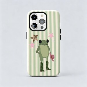 Puede incluir: Una funda para smartphone con fondo de rayas verticales en verde claro y crema. El diseño incluye una ilustración central de una rana verde con botas de color verde oscuro con detalles de corazones, sosteniendo un sombrero rosa, rodeada de estrellas rosas y de leopardo.