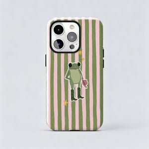 May include: A smartphone case featuring a vertical striped pattern in light pink and olive green. Centered on the case is an illustration of a green frog wearing dark green boots with heart details and holding a pink hat, surrounded by small yellow stars.