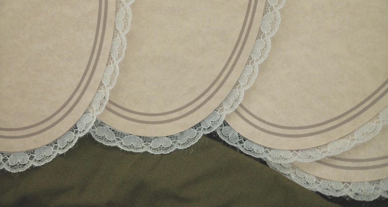 May include: Close-up of several beige placemats with a scalloped white lace trim. Each placemat has two thin, parallel gray lines near the edge. The background is a dark olive green fabric.