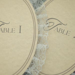 May include: Two oval-shaped table cards with the text "Table 1" and "Table 2" in a script font. The cards are beige with a double brown border and are separated by a white lace trim. The cards are likely for a wedding or event.