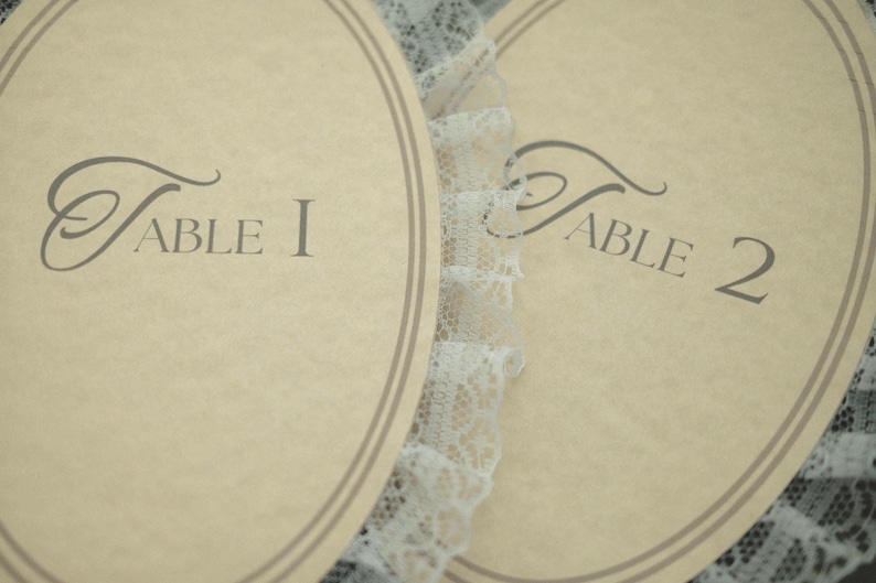 May include: Two oval-shaped table cards with the text "Table 1" and "Table 2" in a script font. The cards are beige with a double brown border and are separated by a white lace trim. The cards are likely for a wedding or event.