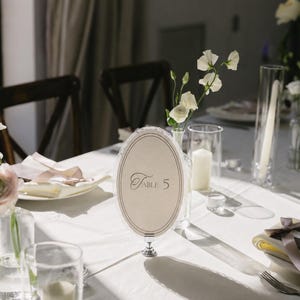 May include: An elegant table setting with a white tablecloth, featuring a decorative oval table number card that reads "Table 5". The scene includes floral arrangements, candles, and glassware, creating a sophisticated ambiance.