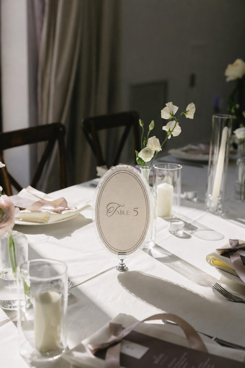 May include: An elegant table setting with a white tablecloth, featuring a decorative oval table number card that reads "Table 5". The scene includes floral arrangements, candles, and glassware, creating a sophisticated ambiance.