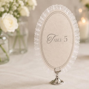 May include: An oval table number card with a delicate lace trim and silver base. The card displays the number "5" in elegant script. Soft white roses and candles create a romantic ambiance, perfect for a wedding or special event.