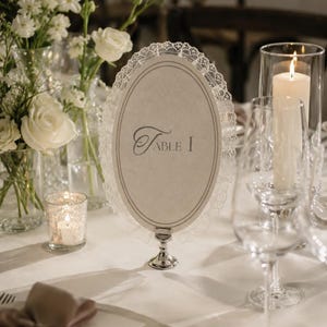 Może przedstawiać: Owalna karta z numerem stołu z koronkową obwódką, z napisem "Table I" w eleganckiej czcionce. Karta umieszczona jest na srebrnym stojaku, otoczona białymi różami, świecami i szkłem, tworząc wyrafinowaną atmosferę.
