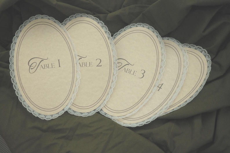 May include: Five oval table number cards with a cream-colored background and a delicate lace trim. Each card is labeled with a table number from 1 to 4 in elegant script. The cards are arranged on a dark green fabric.