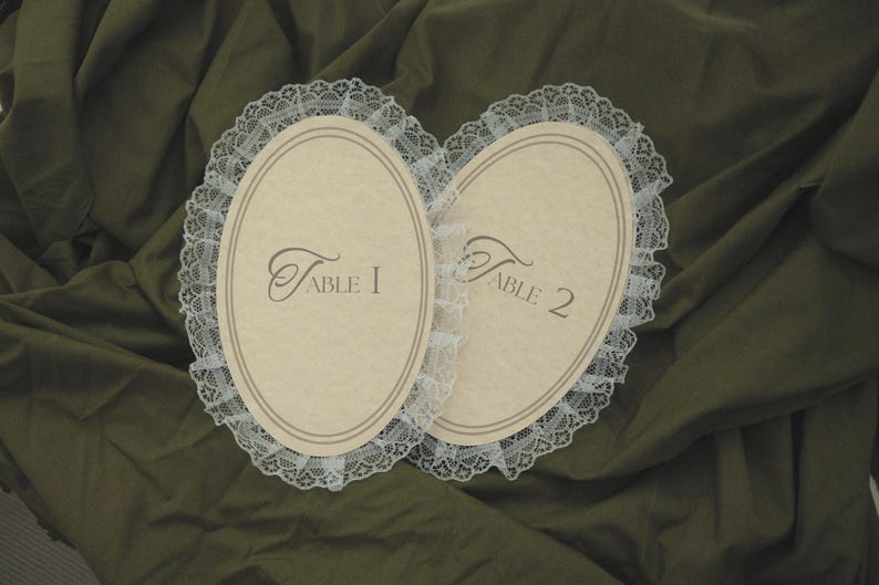 May include: Two oval-shaped table cards with a cream-colored background and a delicate white lace trim. The cards are labeled "Table 1" and "Table 2" in elegant script, perfect for wedding or event decor.