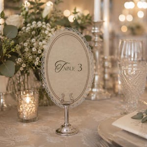 May include: An elegant table setting with a floral centerpiece, candles, and a table number. The table number is an oval card with the text "Table 3" in a decorative font, held by a silver stand. The setting includes glassware and a folded napkin.