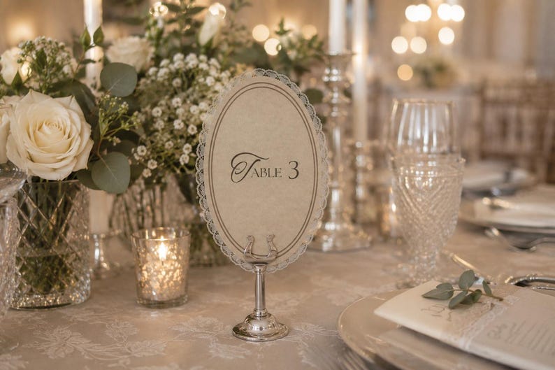 May include: An elegant table setting with a floral centerpiece, candles, and a table number. The table number is an oval card with the text "Table 3" in a decorative font, held by a silver stand. The setting includes glassware and a folded napkin.