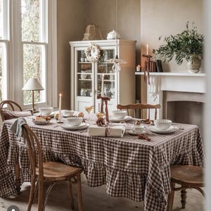 Peut inclure: Une nappe à carreaux marron et blanc avec un bord à volants drapée sur une table à manger. La table est dressée avec des bols et des assiettes en céramique blanche cannelée, ainsi qu'un cadeau emballé avec un ruban marron.