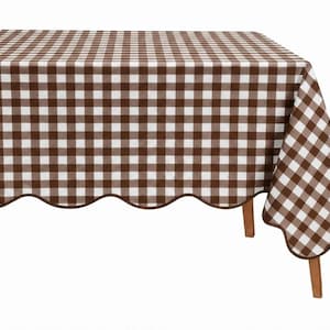 May include: A rectangular tablecloth featuring a brown and white gingham check pattern. The fabric has a scalloped edge finished with a thin brown trim, draped over a table with visible wooden legs on a plain white background.