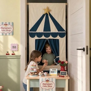 Puede incluir: Una cortina de tela para puerta con diseño de carpa de circo en azul y crema con una estrella amarilla. Una mesa de juego de madera al frente muestra un soporte para cupcakes, una caja registradora de juguete y un letrero Mariana's Cake Shop.
