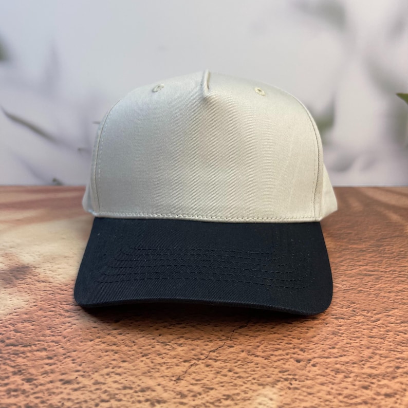 May include: A baseball cap with a two-tone design. The top of the cap is a light beige color, while the brim is a dark navy blue. The cap is made of a smooth fabric and has a classic, structured shape. This hat is suitable for casual wear.