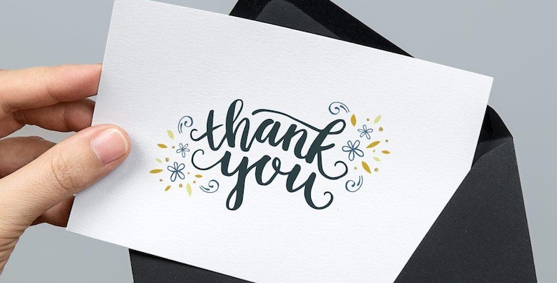 May include: A white rectangular greeting card featuring the dark blue cursive text thank you, surrounded by small blue flower illustrations and gold leaf accents, partially inserted into a solid black envelope with a pointed flap.