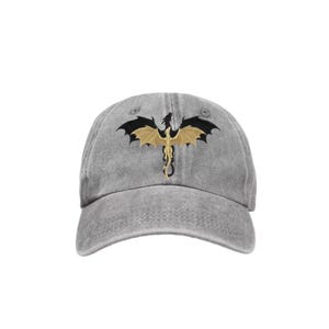 May include: A gray baseball cap featuring a gold and black dragon design. The dragon is embroidered on the front of the cap, with its wings spread wide. The cap has a curved brim and an adjustable strap.