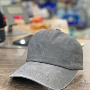May include: A gray baseball cap with a curved brim. The cap is made of a soft fabric and has a classic, unstructured design. The brim features a row of stitching.