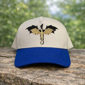 May include: A beige baseball cap with a blue brim. The cap features an embroidered dragon design in black and gold. The dragon is centered on the front of the cap, with its wings spread wide. The cap is sitting on a stone surface.