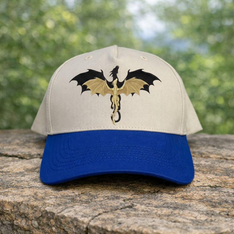 May include: A beige baseball cap with a blue brim. The cap features an embroidered dragon design in black and gold. The dragon is centered on the front of the cap, with its wings spread wide. The cap is sitting on a stone surface.