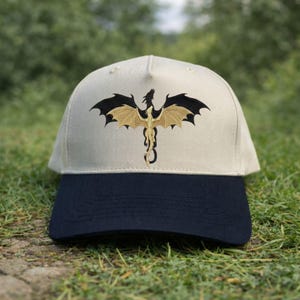 May include: A beige baseball cap with a navy blue brim. The cap features an embroidered dragon design in black and gold. The dragon is centered on the front of the cap, with its wings spread wide. The cap is sitting on green grass.