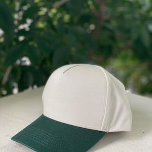 May include: A baseball cap with a white crown and a dark green brim. The cap is made of a sturdy fabric and has a classic, structured design. The background is a blurred view of green foliage.