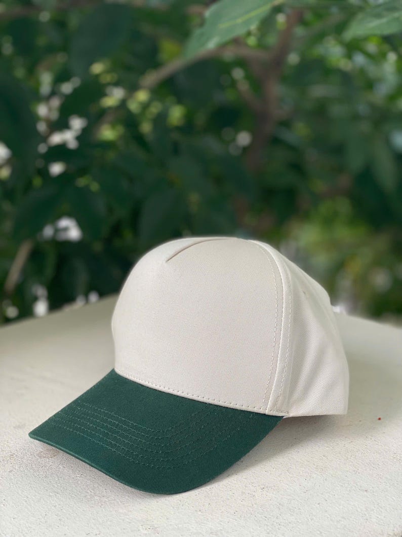May include: A baseball cap with a white crown and a dark green brim. The cap is made of a sturdy fabric and has a classic, structured design. The background is a blurred view of green foliage.