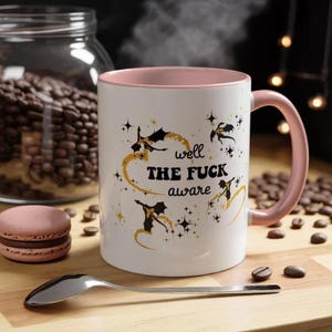 May include: A white ceramic mug with a soft pink interior and handle. The exterior features black silhouettes of dragons, gold swirling accents, and the text "well THE FUCK aware" printed in black, surrounded by small black and gold star icons.