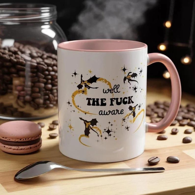 May include: A white ceramic mug with a soft pink interior and handle. The exterior features black silhouettes of dragons, gold swirling accents, and the text "well THE FUCK aware" printed in black, surrounded by small black and gold star icons.