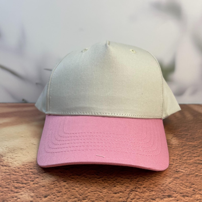 May include: A baseball cap with a two-tone design. The top of the cap is a light beige color, while the brim is a solid pink. The cap is made of a durable fabric and features a curved brim.