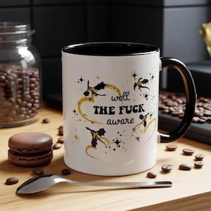 May include: A white ceramic mug with a black interior and handle, featuring black and gold dragon silhouettes, star accents, and the text "well THE FUCK aware" printed in the center. The mug is positioned on a wooden surface with coffee beans.