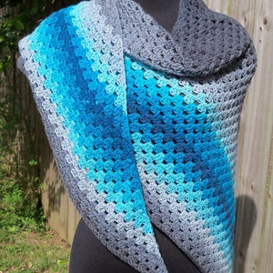 Pode incluir: Um xale triangular de crochê em tons de cinza e azul. O xale está drapeado sobre um manequim e tem uma borda franjada.