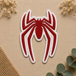 May include: A red and white embroidered patch featuring a stylized spider emblem. The design consists of a central red spider body with long, angular legs extending outward, all outlined with a thick white border on a textured fabric base.