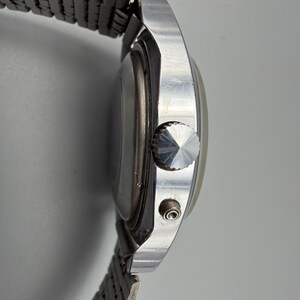 May include: Side view of a silver-toned wristwatch with a metal band. The watch face is round, and the band is made of small, connected metal links. The watch has a textured crown and a small button on the side.