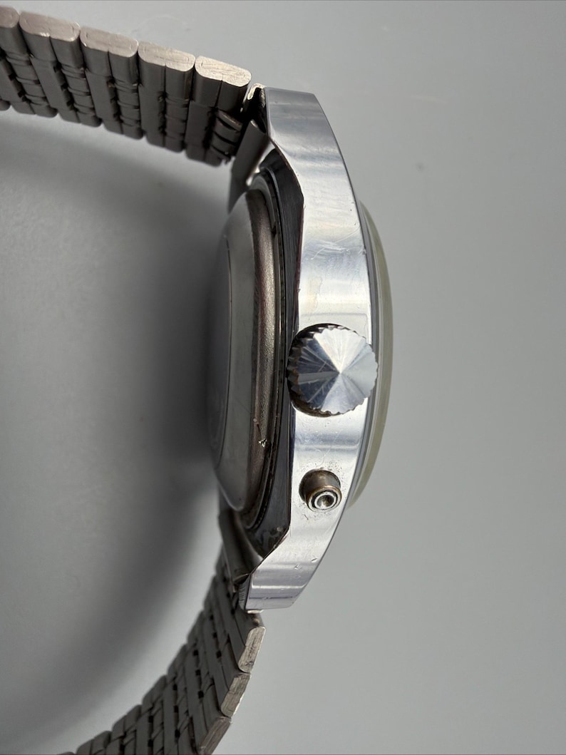 May include: Side view of a silver-toned wristwatch with a metal band. The watch face is round, and the band is made of small, connected metal links. The watch has a textured crown and a small button on the side.