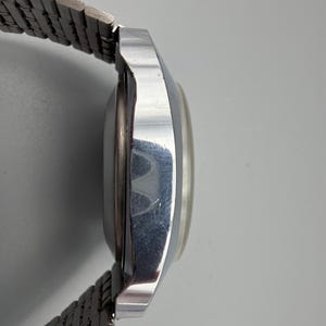 May include: Side view of a vintage silver-toned wristwatch with a metal band. The watch face is round and has a light-colored dial. The band is made of small, interconnected metal links.