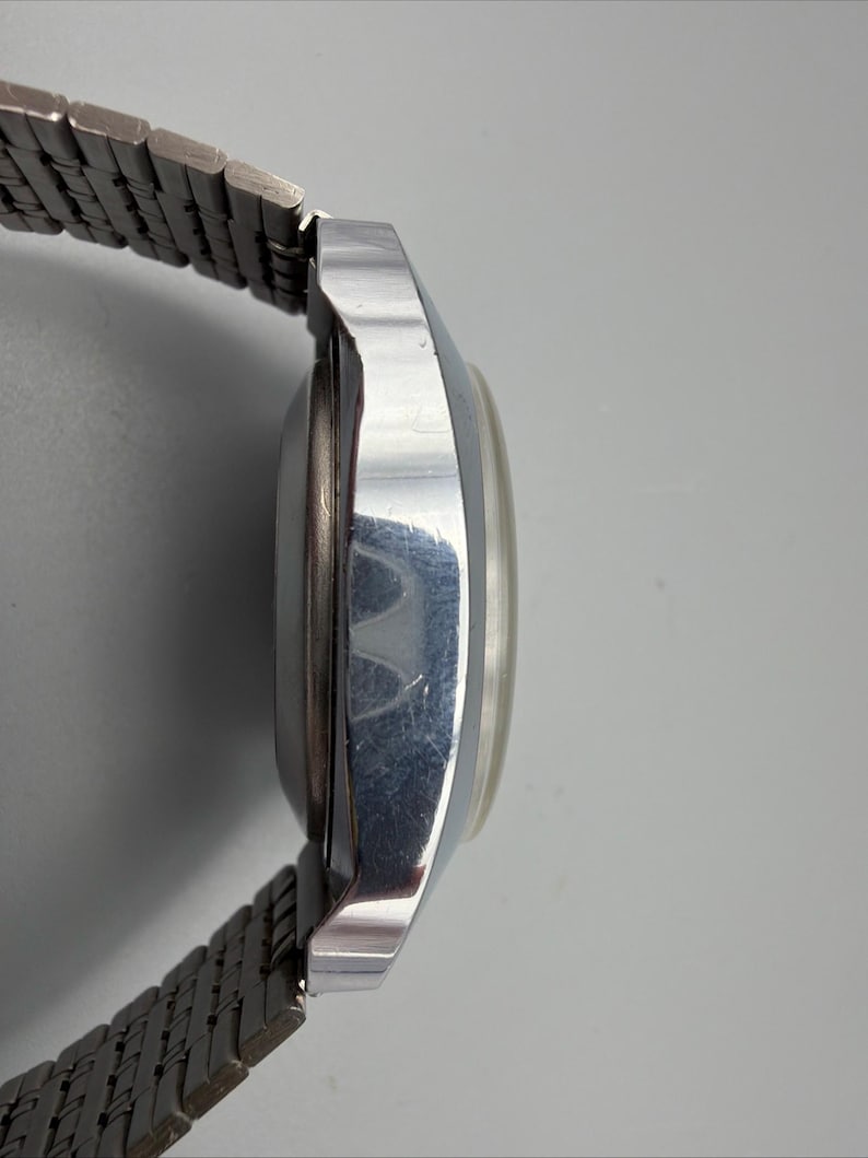 May include: Side view of a vintage silver-toned wristwatch with a metal band. The watch face is round and has a light-colored dial. The band is made of small, interconnected metal links.