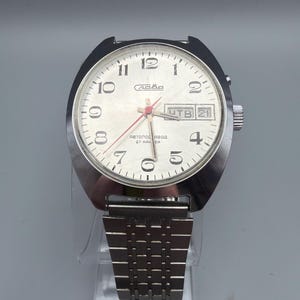 May include: A vintage silver-toned wristwatch with a white face, black numerals, and red hands. The watch features a day and date display and a stainless steel band. The watch face has the word "Слава" and other text.