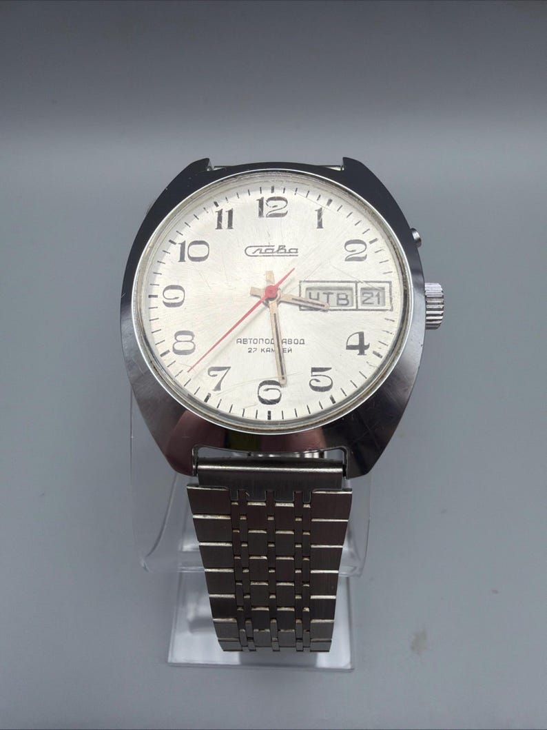 May include: A vintage silver-toned wristwatch with a white face, black numerals, and red hands. The watch features a day and date display and a stainless steel band. The watch face has the word "Слава" and other text.