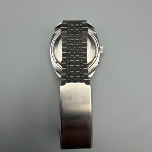 May include: A vintage silver-toned wristwatch with a rectangular face and a linked metal band. The watch has a brushed metal finish and a clasp closure. The watch is displayed against a gray background.