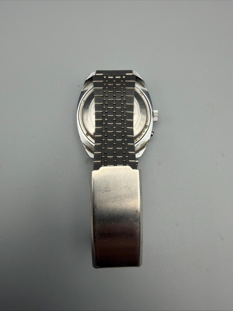 May include: A vintage silver-toned wristwatch with a rectangular face and a linked metal band. The watch has a brushed metal finish and a clasp closure. The watch is displayed against a gray background.