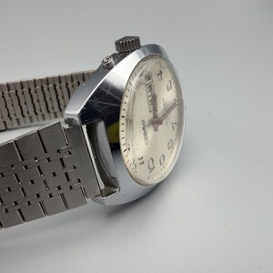 May include: Silver-toned wristwatch with a light-colored face and a metal band. The watch face displays numbers and hands, with a day and date display. The band is made of interlocking metal links. The watch is angled, showing the side and face.