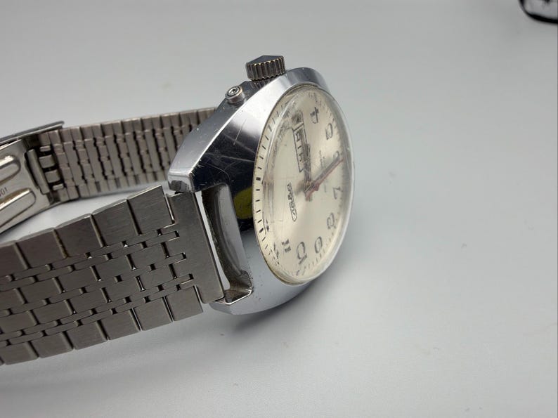 May include: Silver-toned wristwatch with a light-colored face and a metal band. The watch face displays numbers and hands, with a day and date display. The band is made of interlocking metal links. The watch is angled, showing the side and face.