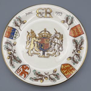 Könnte beinhalten: Eine weiße Gedenkplatte mit goldenem Rand, mit dem königlichen Wappen, Eichenblättern und Eicheln. Die Platte trägt den Text "June 1953" und die Initialen "ER" in Gold.