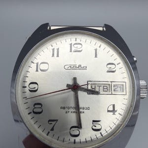 May include: Silver-toned wristwatch with a round face and a silver band. The watch face has black numerals, a day-date display, and the word "Слава" in Cyrillic script. The watch hands are red.